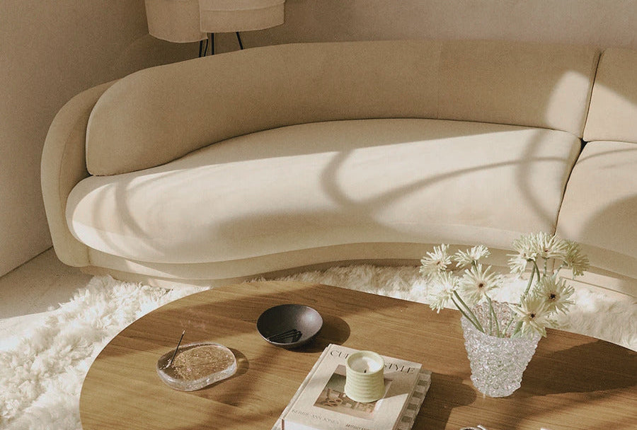 A modern curved beige sofa in a warm, minimalist living room setting with a round wooden coffee table, a fluffy white rug, and soft natural sunlight casting shadows.